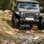 A black Jeep Wrangler is driving through a shallow creek filled with rocks. There are trees on both sides of the vehicle.