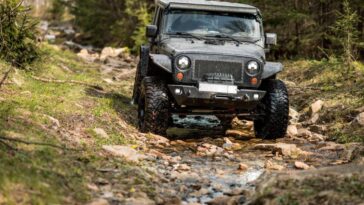 A black Jeep Wrangler is driving through a shallow creek filled with rocks. There are trees on both sides of the vehicle.