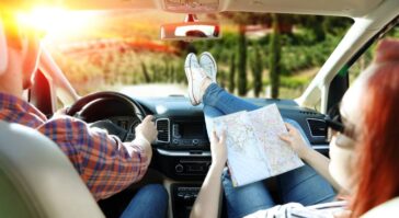 A man in a plaid shirt is driving an SUV into a sunset. A woman with her feet up on the dash is looking over a map.