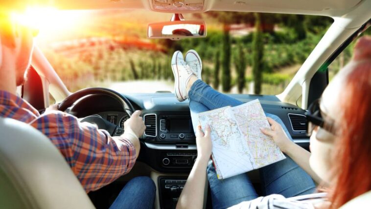 A man in a plaid shirt is driving an SUV into a sunset. A woman with her feet up on the dash is looking over a map.