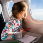 A young girl is sitting on an airplane and looking out the window. She is drawing a picture with colored pencils.