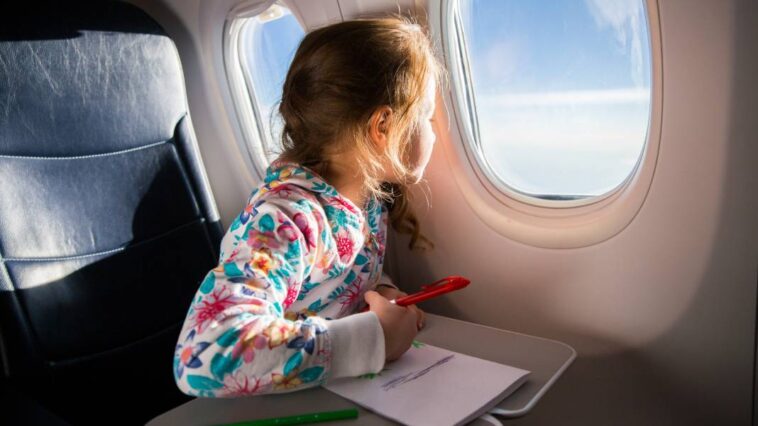 A young girl is sitting on an airplane and looking out the window. She is drawing a picture with colored pencils.