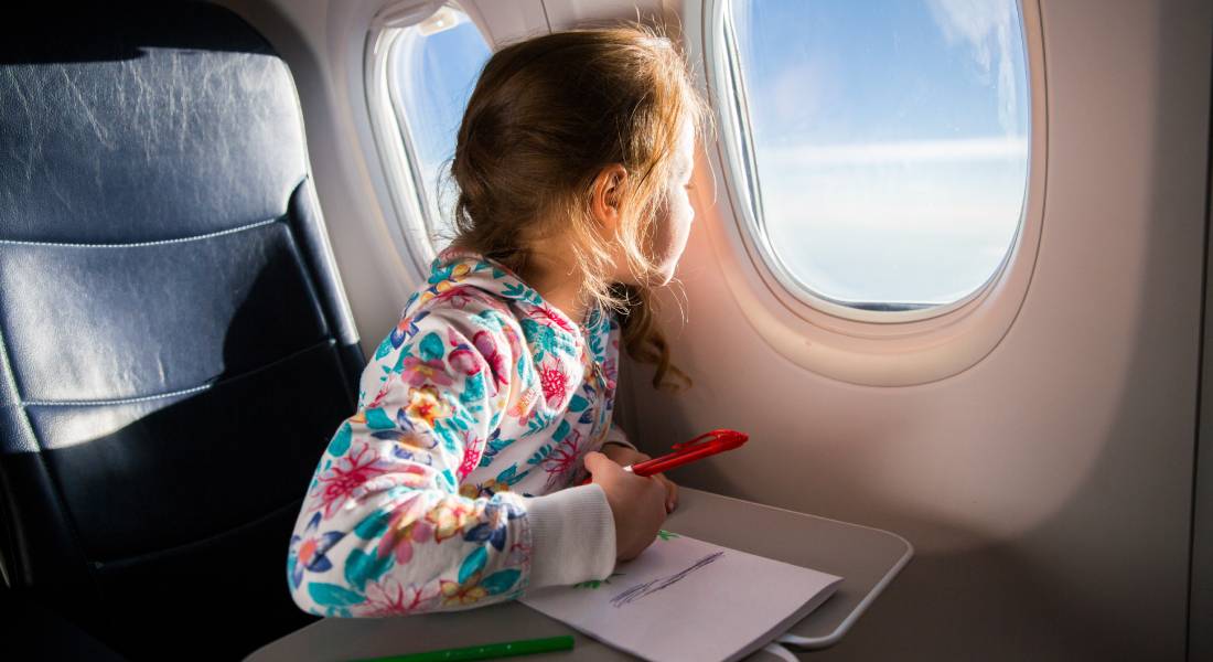 A young girl is sitting on an airplane and looking out the window. She is drawing a picture with colored pencils.