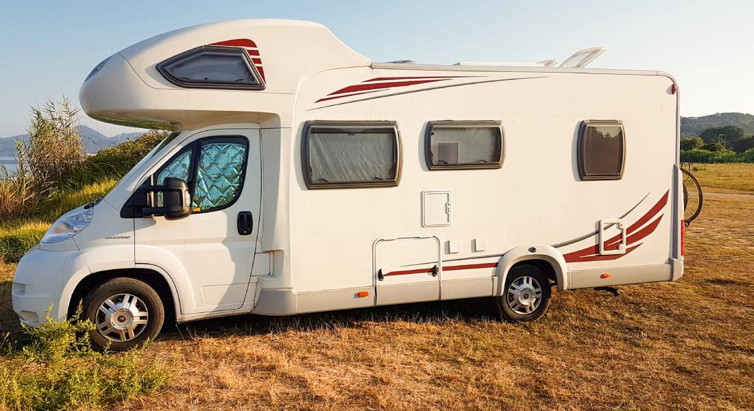 A white RV is parked outside in a field on a sunny day. The windshield and side windows are covered by sunshades.